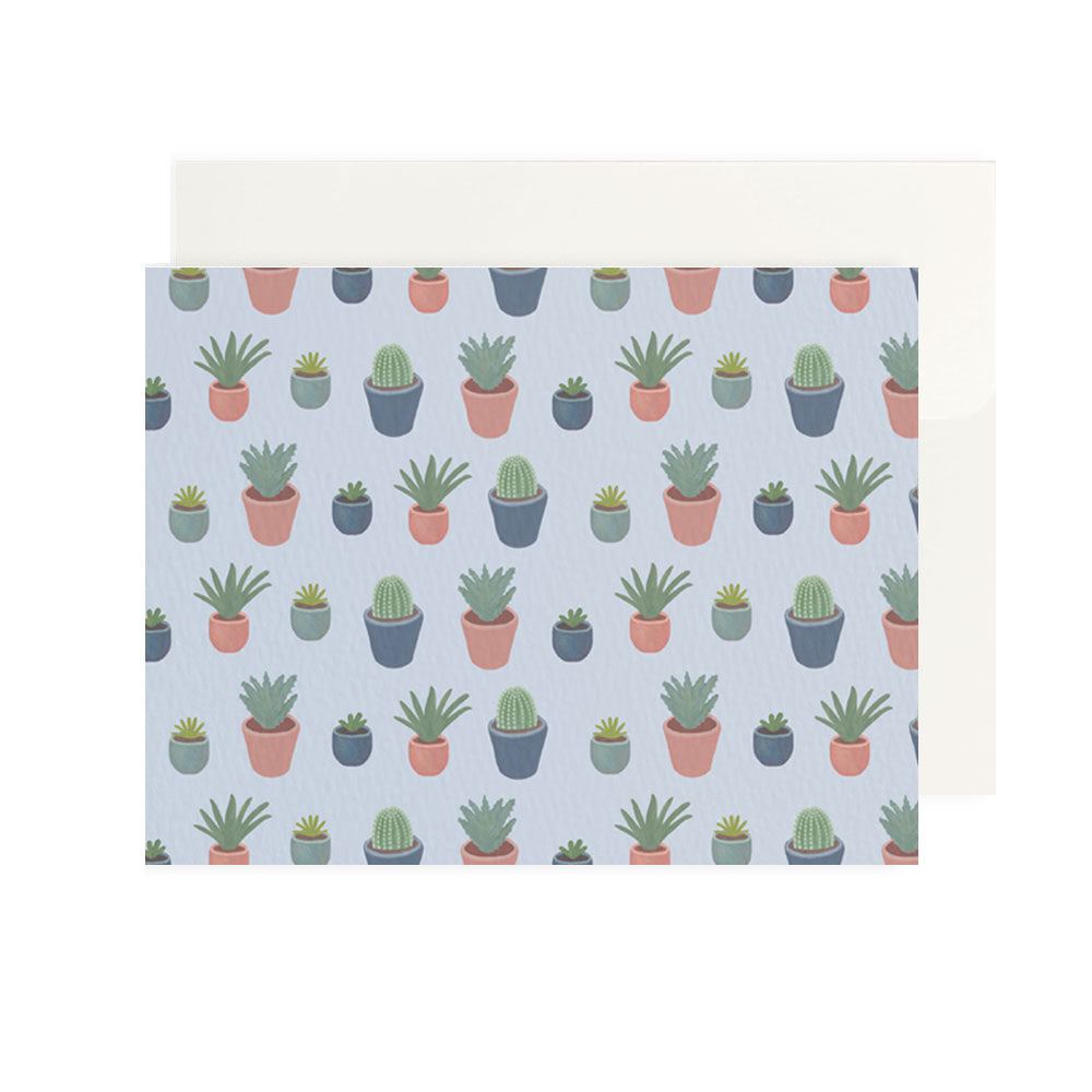 A flat, front-facing view of a horizontal greeting card placed in front of a plain off-white envelope. The card features a repeating pattern of various illustrated green cacti and succulents in small pots colored terracotta pink, muted teal, and dark grey. The entire pattern is set against a soft, pale lavender-blue background.