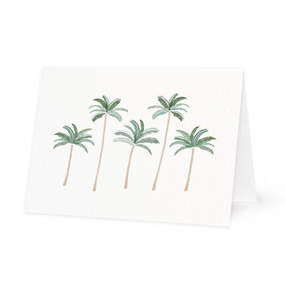 a horizontal folded greeting card standing slightly open on a white surface. the front of the card is made of textured white paper and features a watercolor illustration of five palm trees with green fronds and tall, slender tan trunks. the background is plain white.