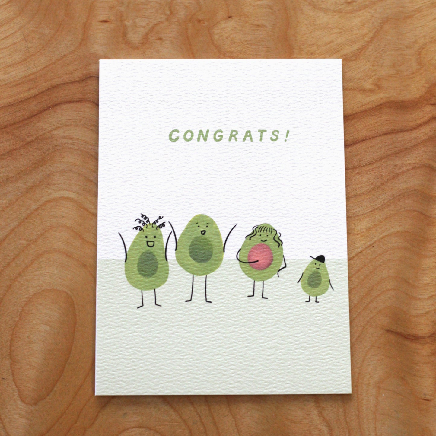 A vertical white greeting card featuring four illustrated avocado cartoon characters standing in a row on a light green ground. From left to right: an avocado with curly hair and arms raised, a tall avocado with a wide smile, a pregnant avocado with an avocado seed belly, and a small avocado wearing a black hat. The word 'Congrats!' is printed in light green sans-serif capital letters above them. the card lays flat on a wood surface.