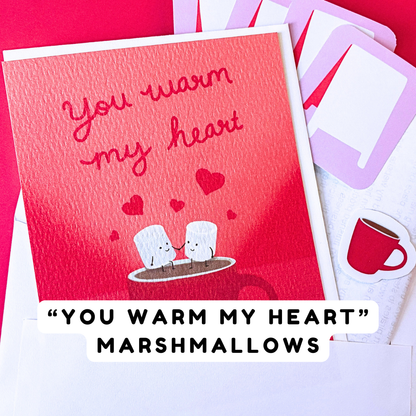 A red greeting card with the phrase “you warm my heart” in cursive script, decorated with two cartoon marshmallows holding hands sitting on a rim of a red mug with steam and background red hearts. Partially visible behind it are four sticker labels with a-frame sidewalk sign design and a red mug sticker. There is a white text banner reading “You Warm My Heart Marshmallows".
